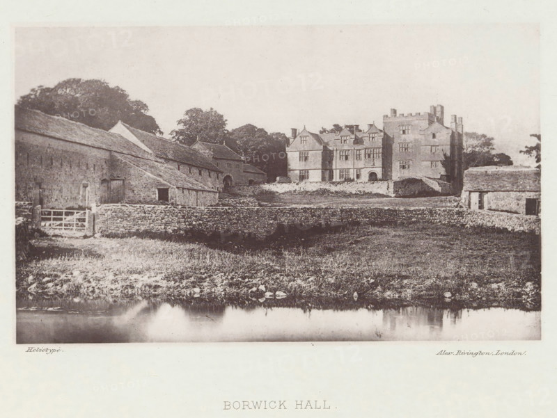 Borwick Hall, View of Borwick Hall, page, photomechanical print, photographer: Alex Rivington, Borwick, London, c. 1870 - in or before 1880, paper, heliotype, height 106 mm x width 175 mm, rural housing, eg country-house, villa, cottage, Lancashire, A wide sepia-toned view shows a large stone manor house with multiple gabled roofs and tall chimneys set behind a low stone wall, flanked by adjoining stone outbuildings with pitched roofs and small doorways; a grassy foreground with scattered rocks slopes down to a reflective body of water at the bottom edge, and a cluster of trees rises behind and between the buildings.