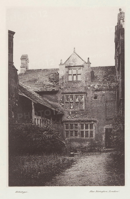 Borwick Hall, View of Borwick Hall, page, photomechanical print, photographer: Alex Rivington, Borwick, London, c. 1870 - in or before 1880, paper, heliotype, height 179 mm x width 117 mm, rural housing, eg country-house, villa, cottage, Lancashire, A tall stone building facade with a pitched roof and multiple leaded windows set in mullioned frames, showing aged and weathered textured walls and roof tiles; a covered wooden gallery or porch projects from the left side with visible timber posts and beams, and dense foliage or shrubs grow along the base of the building beneath the gallery; a gravel or dirt path runs toward the foreground from the building entrance area, and parts of adjacent stone walls and chimneys are visible at the image edges.