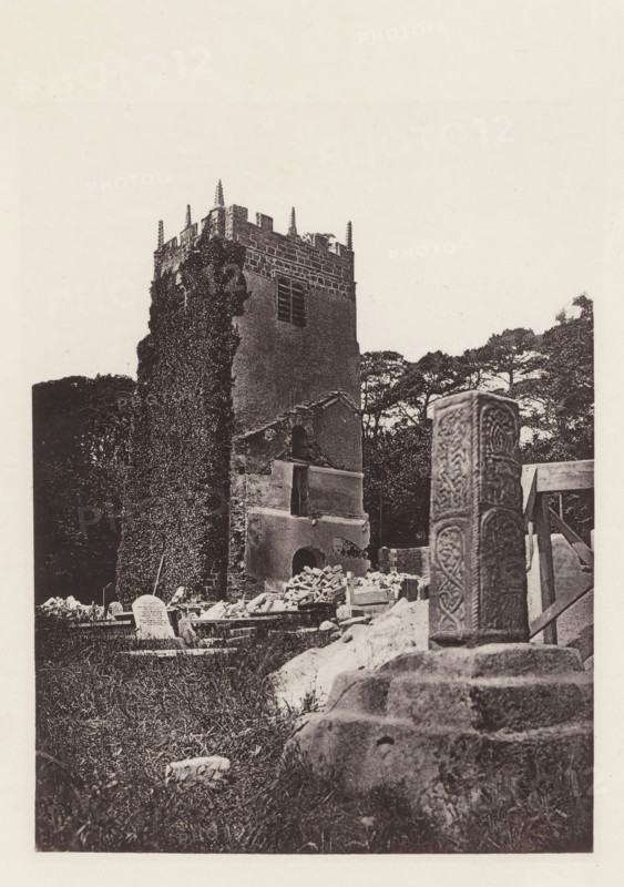 Tower of a church in Halton-with-Aughton, Halton church tower. 1876, page, photomechanical print, photographer: Wright, HM & Co., Halton-with-Aughton, London, 1876, paper, heliotype, height 150 mm x width 112 mm, parts of church exterior and annexes: tower, Lancashire, A tall stone tower with battlements and small pointed pinnacles rises behind a partially ruined, ivy-covered wall, adjacent to scattered masonry and broken architectural elements; in front of the tower a carved upright stone pillar with ornate interlaced patterns stands on weathered steps, and gravestones and rubble lie across a grassy foreground with trees visible behind the tower.