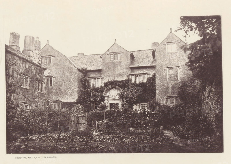Cark Hall, View of a country house in Cark, page, photomechanical print, photographer: Alex Rivington, Cark, London, c. 1870 - c. 1880, paper, heliotype, height 114 mm x width 180 mm, rural housing, eg country-house, villa, cottage, Cumbria, A sepia-toned photograph of an old multi-story building with textured exterior walls and steeply pitched roofs, featuring dormer windows and tall chimneys; ivy and climbing plants cover portions of the facade and surround a central ornate arched doorway with decorative stonework, while rectangular windows of varying sizes punctuate the walls; in front of the building a garden area with rows of low plants, trellises, and a small wooden gate is visible, and dense foliage frames the right side of the scene.