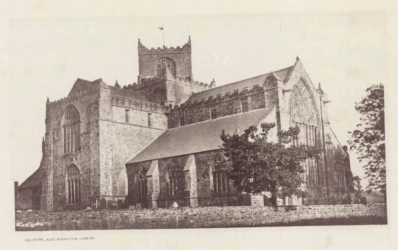 View of the Cartmel Priory, Cartmel church, page, photomechanical print, photographer: Alex Rivington, Cartmel, London, c. 1870 - in or before 1880, paper, heliotype, height 104 mm x width 180 mm, church (exterior), Lancashire, Cartmel Priory, A large stone building with tall arched windows and a peaked roof dominates the image, showing detailed masonry, buttresses, and crenellated parapets; a square tower with a louvered opening and a flagpole rises above the central roofline; trees partially obscure the lower right portion of the structure; a low stone wall runs along the foreground.