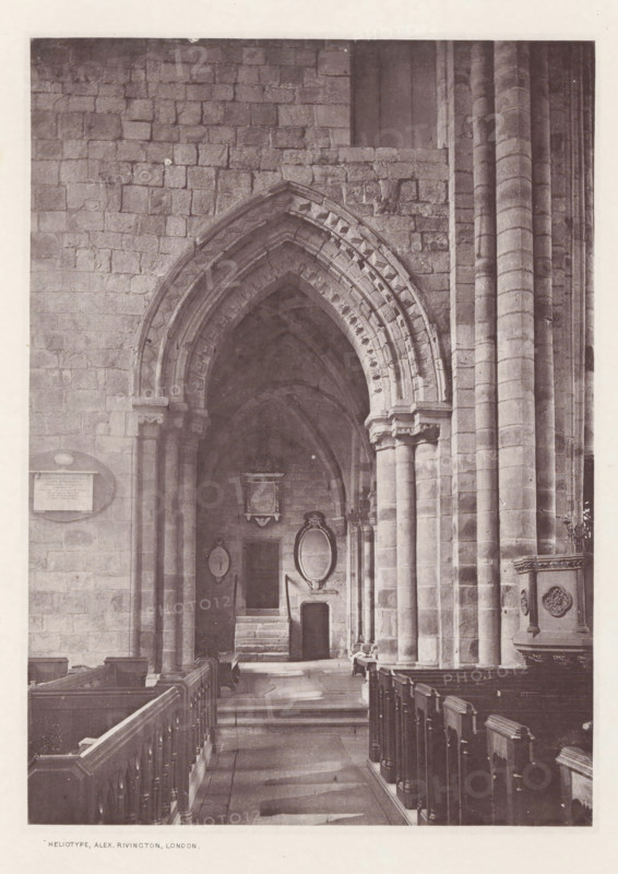 Piper choir. Cartmel church, Interior of the Cartmel Priory, page, photomechanical print, photographer: Alex Rivington, Cartmel Priory, London, c. 1870 - in or before 1880, paper, heliotype, height 177 mm x width 128 mm, interior of church, parts of church interior: choir, Lancashire, Cartmel, A stone interior of a building showing a long aisle with wooden pews on either side leading toward an arched doorway; the walls and arch are constructed from large rectangular blocks and the arch features layered, decorative mouldings. Tall cylindrical columns rise along the right side supporting the structure, and a carved stone pulpit with decorative panels is visible near the foreground on the right. Within the archway there are stone steps, recessed niches, and framed oval and rectangular plaques mounted on the wall, with a small door set beneath one of the plaques. The floor shows worn stone paving and shadow patterns cast by the architecture.