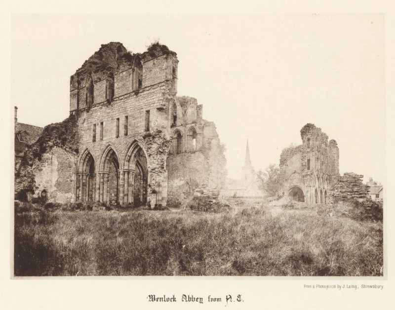 Wenlock Abbey from NE, View of the ruins of Wenlock Priory, page, photomechanical print, photographer: J. Laing, Wenlock Priory, cliché maker: anonymous, c. 1867 - in or before 1877, Shropshire, Much Wenlock, paper, collotype, photoprint, height 137 mm x width 193 mm, ruin of church, monastery, A wide, sepia-toned scene shows the ruins of a large stone abbey with tall, arched openings and partially collapsed walls, vegetation growing on and around the masonry, an overgrown grassy foreground, scattered remnants of arches and window openings, a distant slender spire rising behind the ruins, and a few small buildings partially visible near the edges of the composition.