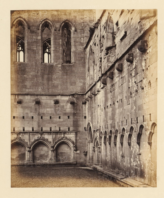 Remains of the cloister of Melrose Abbey, photograph, page, inscription: 'With finely-carved Door on the right, ', number: '(No. 9.)', title: 'Remains of ancient cloisters', photographer: WJ Pringle, Melrose Abbey, c. 1860 - in or before 1870, Melrose (Scotland), paper, albumen print, height 171 mm x width 140 mm, ruin of church, monastery, cloisters, A sepia-toned photograph of a stone building corner showing two façades with Gothic architectural details, including tall, narrow arched windows with tracery on the left wall and a row of smaller blind arches along the right wall. The left façade features large rectangular stone blocks and high pointed windows set into deep reveals, while the lower section shows a series of ornate pointed arches resting on clustered columns. The right façade displays weathered stone, projecting corbels near the top, repeated blind arches with molded surrounds, patches of lighter discoloration on the surface, and a narrow ledge running along the base. The ...