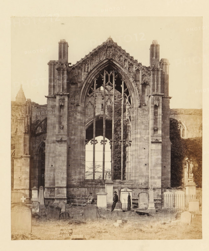 View of a window of the ruins of Melrose Abbey with two men in front of it, photograph, page, title: 'East window', number: '(No. 11.)', inscription: 'Thirty-seven feet in height by sixteen in breadth, at the point of the arch are figures of David I., the Founder of the Abbey, and Matilda, his Queen.', photographer: WJ Pringle, Melrose Abbey, c. 1860 - in or before 1870, Melrose (Scotland), paper, albumen print, height 164 mm x width 141 mm, ruin of church, monastery, church-window, eg rose-window, A sepia-toned photograph of a large Gothic stone façade with a tall pointed arched window featuring ornate tracery and vertical stone mullions, flanked by two tall buttresses and decorative pinnacles; the lower portion shows a graveyard with various upright and leaning headstones, a fenced area, and two men among the monuments, one seated on a bench and the other standing and leaning against a gravestone; ivy or climbing vegetation is visible along part of the right side of the façade and sm...