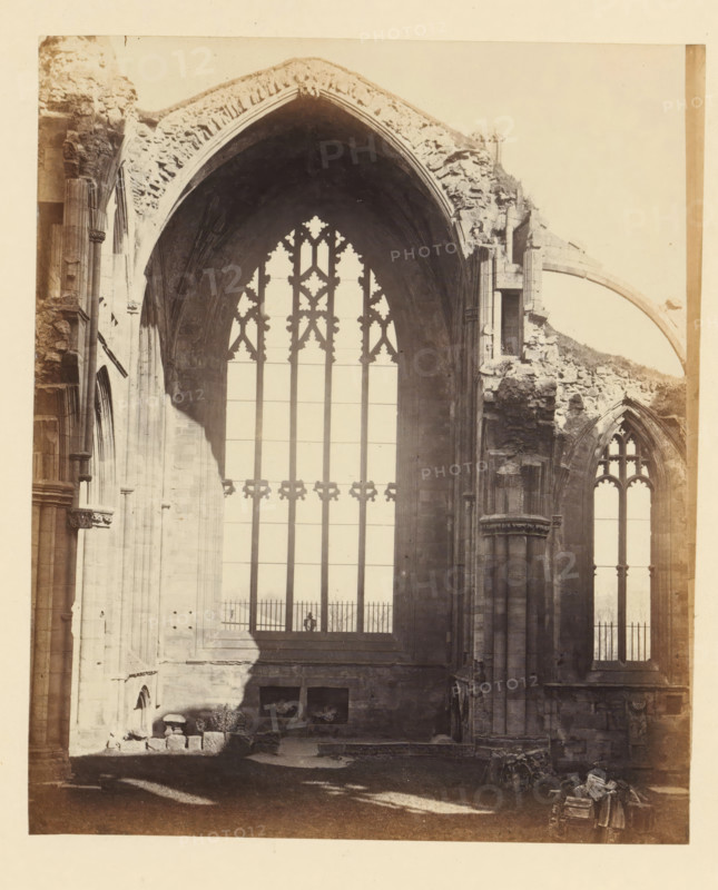View of a window of the ruins of Melrose Abbey, photograph, page, number: '(No. 12.)', title: 'Interior', inscription: 'Exhibiting the Chancel, and situation of High Altar, with Tomb of Alexander II. of Scotland, and that in wich the Heart of Robert Bruce was deposited.', photographer: WJ Pringle, Melrose Abbey, c. 1860 - in or before 1870, Melrose (Scotland), paper, albumen print, height 165 mm x width 137 mm, ruin of church, monastery, church-window, eg rose-window, A sepia-toned photograph showing the interior of a large stone building with tall arched window openings and ornate tracery, with one central high window framed by a pointed arch and flanked by columns and ruined masonry; portions of the stone walls and archways show weathered and broken edges, rubble and scattered stones lie on the floor near the base of the walls, a shadow falls across part of the wall and floor, and a smaller arched window is visible to the right with Gothic tracery.