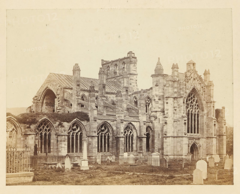 View of the ruins of Melrose Abbey, photograph, page, number: '(No. 1.)', title: 'The Abbey from the south-west', photographer: WJ Pringle, Melrose Abbey, c. 1860 - in or before 1870, Melrose (Scotland), paper, albumen print, height 134 mm x width 173 mm, ruin of church, monastery, A sepia-toned photograph of a large stone Gothic-style building with arched windows and ornate tracery, sections of the façade showing pointed arches and tall narrow windows, buttresses and decorative pinnacles, and a partially ruined archway on one side; in front of the building there is a grassy area with fenced enclosures, stone tombstones and monuments including an obelisk, and wrought iron railings surrounding some graves.