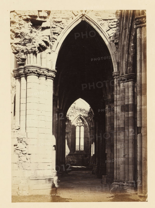 View of an aisle of the ruins of Melrose Abbey, photograph, page, title: 'Fine perspective view of south aisle', number: '(No. 4.)', inscription: 'The Carving and Capitals of Pillars, remarkable', photographer: WJ Pringle, Melrose Abbey, c. 1860 - in or before 1870, Melrose (Scotland), paper, albumen print, height 171 mm x width 121 mm, ruin of church, monastery, parts of church interior: aisle, A tall stone archway framed by thick columns and ornate capitals leads into a dark vaulted interior with layered arches receding toward a distant pointed window, rough stone walls and carved column shafts visible on either side, light filtering through the far window creating contrasts of shadow and pale stone on the floor and lower walls.