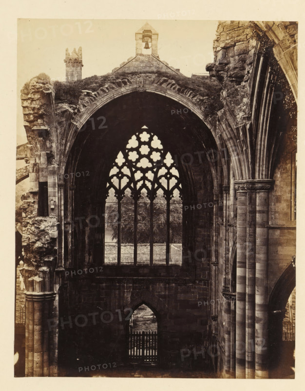 Interior with a view of a window of the ruins of Melrose Abbey, photograph, page, number: '(No. 5.)', inscription: 'With Belfry and Font to the right.', title: 'Interior of south transept', photographer: WJ Pringle, Melrose Abbey, c. 1860 - in or before 1870, Melrose (Scotland), paper, albumen print, height 175 mm x width 138 mm, ruin of church, monastery, church-window, eg rose-window, A tall stone arch with a tracery window sits between massive ribbed columns and weathered masonry, the window opening filled with ornate quatrefoil and trefoil shapes revealing trees and a fence beyond, rough stonework and carved capitals flank the arch on both sides, an arched doorway with a gate is visible below, broken and crumbling sections of the structure show exposed layers of stone and patches of vegetation, and a small bell tower rises above the archline against a pale sky.