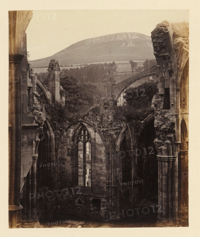View of a chapel of the ruins of Melrose Abbey with Eildon Hill in the background, photograph, page, title: 'Chapel to east of south transept', number: '(No. 6.)', inscription: 'With Tomb of Michael Scott, the Wizard, and Eildon Hill in the distance.', photographer: WJ Pringle, Melrose Abbey, c. 1860 - in or before 1870, Melrose (Scotland), paper, albumen print, height 169 mm x width 140 mm, ruin of church, monastery, (high) hill, parts of church interior: chapel, A sepia-toned photograph shows the ruins of a stone building with tall, arched windows and broken, weathered columns and lintels, revealing jagged edges and crumbling masonry; vegetation grows among the ruins and over some upper sections, with leafy trees visible beyond the structure; in the background a gently sloping hill rises behind a patchwork of fields and lines of trees, and an arched stone bridge or buttress spans across part of the scene.