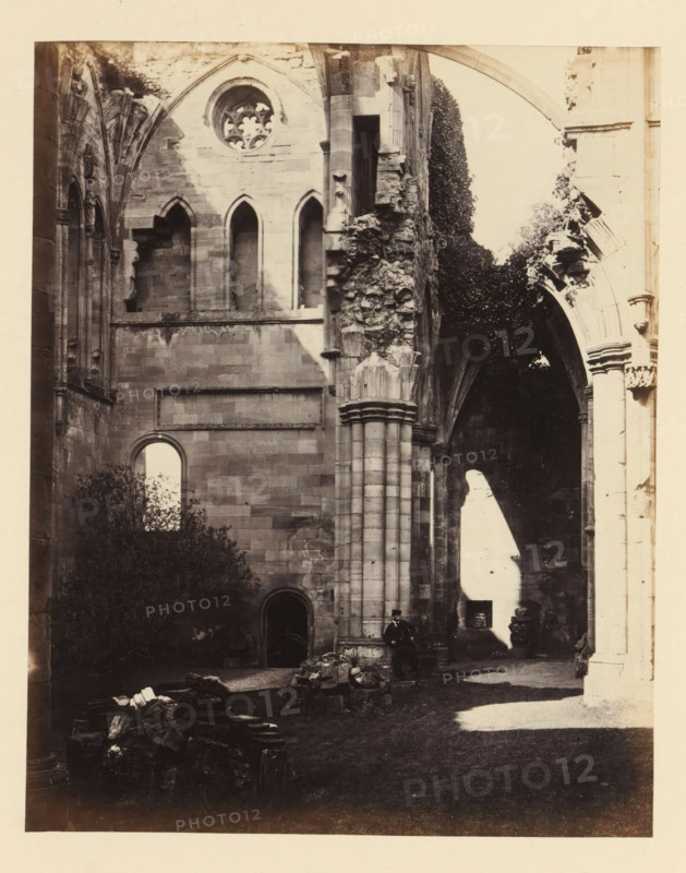 Interior of the north transept of the ruins of Melrose Abbey, photograph, page, title: 'Interior view of north transept', inscription: 'On the left, Statue of St. Peter with the Keys, in front, Crown of Thorns Window.', number: '(No. 7.)', photographer: WJ Pringle, Melrose Abbey, c. 1860 - in or before 1870, Melrose (Scotland), paper, albumen print, height 165 mm x width 131 mm, ruin of church, monastery, parts of church interior: transept, A sepia-toned photograph of a ruined stone building interior showing tall arched openings and a round tracery window high on a wall, with sections of carved columns and crumbling masonry. Ivy or dense foliage grows over part of the ruins near one of the arches. Sunlight creates a bright patch on the floor and a contrasting shadow on the wall. Rubble and scattered stone fragments lie on the ground in the foreground. A man wearing dark clothing and a hat is seated at the base of a large column near the center right of the image.
