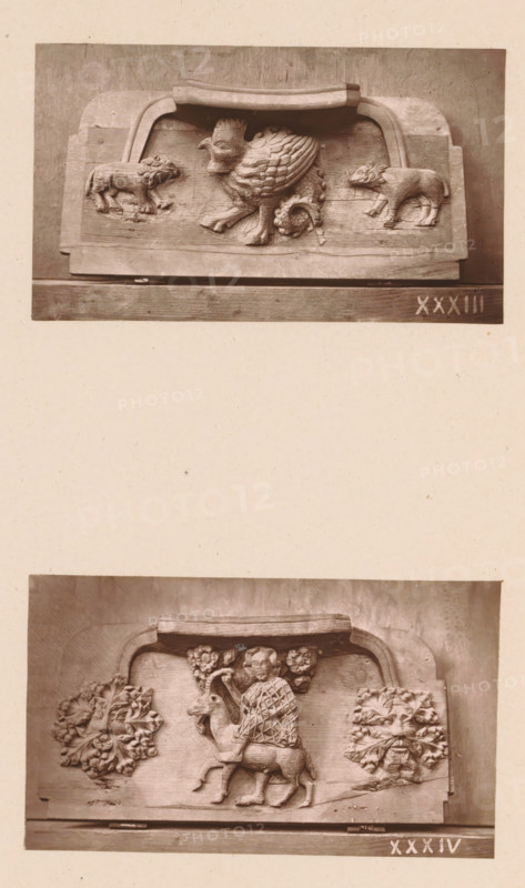 Two woodcarvings in Worcester Cathedral, page, photomechanical print, Above a basilisk between (presumably) two wolves, below a man on a goat between two stylized leaf motifs., number: 'XXXIII, XXXIV', c. 1863 - in or before 1873, paper, Woodburytype, Woodburytypie, height 306 mm x width 244 mm, interior of church, goat, carving, applied arts, basilisk (cock with posterior of snake), A photograph shows two horizontal carved wooden panels mounted one above the other on a pale background, each panel featuring sculpted reliefs within rectangular frames topped by curved caps. The upper panel displays a central griffin-like creature with a scaled body and feathered wings, flanked by two smaller four-legged animals facing inward; the wood shows carved texture and worn surfaces. The lower panel depicts a human figure seated on a stag or similar hoofed animal, holding a curved object, with stylized floral or foliate clusters carved on either side; the figure's garment has a diamond pattern and...