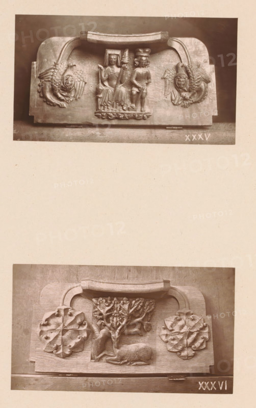 Two wood carvings in Worcester Cathedral, page, photomechanical print, Above a spinning woman and man between two mythical creatures, below a deer between two leaf motifs., number: 'XXXV, XXXVI', c. 1863 - in or before 1873, paper, Woodburytype, height 306 mm x width 244 mm, interior of church, spinning (manufacture of yarn), hoofed animals: stag, carving, applied arts, The image shows two separate rectangular wooden panels photographed one above the other on a light background; the upper panel features a carved central scene of two seated human figures under a curved handle with carved winged faces flanking each side, while the lower panel displays a carved reclining deer beneath a stylized tree with large ornamental leaves on either side and a curved handle above.