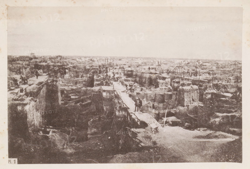 View of a diamond mine in South Africa, presumably near Kimberley, page, photomechanical print, number: 'Pl. 1', photographer: Gray Brothers (possibly), South Africa, c. 1870 - in or before 1880, paper, collotype, photogravure, height 123 mm x width 185 mm, Print in rear image section., mine, Kimberley, A wide sepia-toned photograph showing a rough landscape of deep, irregular trenches and mounds of earth stretching toward the horizon, with narrow plank walkways bridging the gaps between elevated dirt walls and scattered wooden supports and ladders set among the excavated areas; small groups of figures in dark clothing stand or walk along the paths and between the trenches, and various indistinct tools and debris lie on the ground; the sky appears pale and empty above the scene, while the foreground features steep vertical edges of the trenches and rough, textured soil.