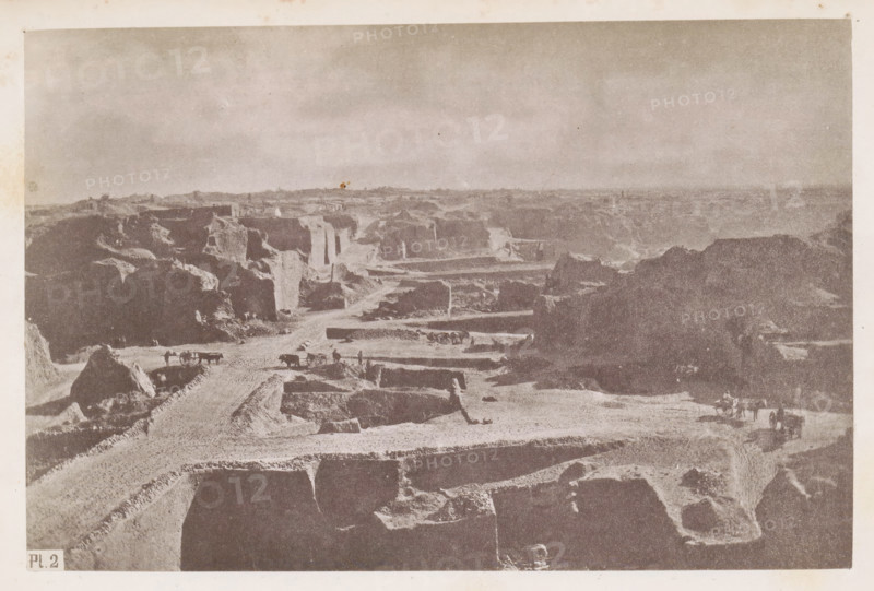 View of a diamond mine in South Africa, presumably near Kimberley, page, photomechanical print, number: 'Pl. 2', photographer: Gray Brothers (possibly), South Africa, c. 1870 - in or before 1880, paper, collotype, photogravure, height 124 mm x width 186 mm, Print in rear image section., mine, Kimberley, A wide, desaturated landscape showing layered, rectangular excavations and steep earthen walls stretching into the distance under a pale sky, with rough dirt roads winding between the cut terraces, scattered large rock formations, several horse-drawn carts and small groups of people on the roads, and distant low ridges along the horizon.