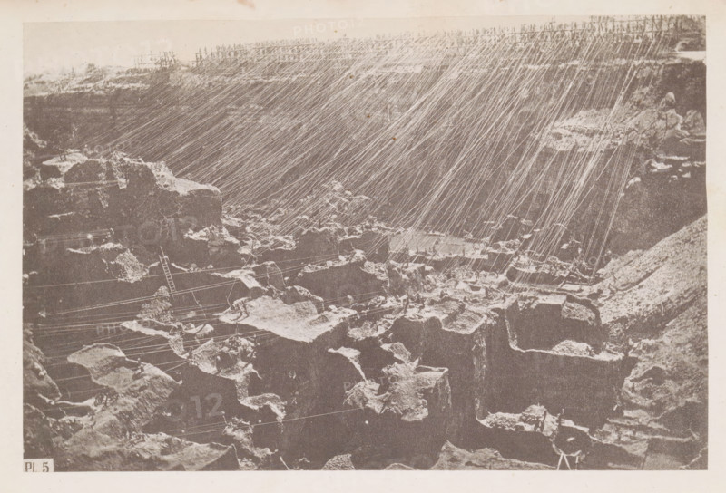 View of the diamond mine at Kimberley in South Africa, page, photomechanical print, number: 'Pl. 5', photographer: Gray Brothers, Kimberley, c. 1870 - in or before 1880, paper, collotype, photogravure, height 126 mm x width 189 mm, Print in rear image section., mine, A sepia-toned photograph of a rocky landscape with steep, jagged cliffs and scattered large boulders, over which numerous thin, bright diagonal lines run from the upper right toward the lower left, creating a streaked pattern across the image; the foreground shows irregular rock surfaces and crevices with lighter patches on some edges, a slender ladder leaning against a rock, and several taut lines stretched horizontally and diagonally among the stones, while the upper edge of the scene reveals a rough rim of rock and a faint horizon.