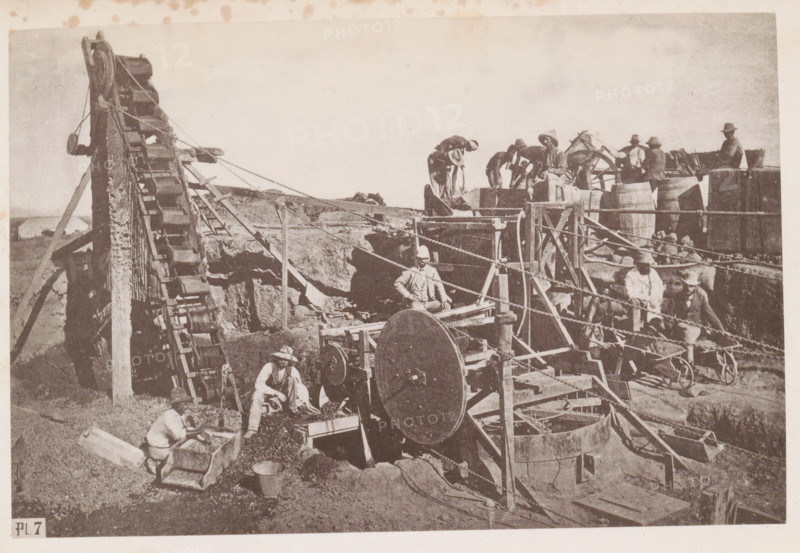 Men at work in a diamond mine in South Africa, presumably near Kimberley, page, photomechanical print, number: 'Pl. 7', photographer: Gray Brothers (possibly), South Africa, c. 1870 - in or before 1880, paper, collotype, photogravure, height 125 mm x width 189 mm, Print in rear image section., mine, Kimberley, A sepia-toned scene shows a group of people working among wooden machinery and barrels in an open, dusty area. A large inclined wooden conveyor with rectangular buckets runs up the left side, supported by a tall frame and ropes. In the center foreground a large round wooden wheel is mounted within a wooden structure with beams and pulleys, and ropes and chains run across the scene connecting various components. Several men wearing wide-brimmed hats and light clothing are positioned around the machinery; some stand on raised platforms near barrels while others crouch or sit at workstations handling material in wooden troughs and carts. Stacked barrels and wooden crates appear near...
