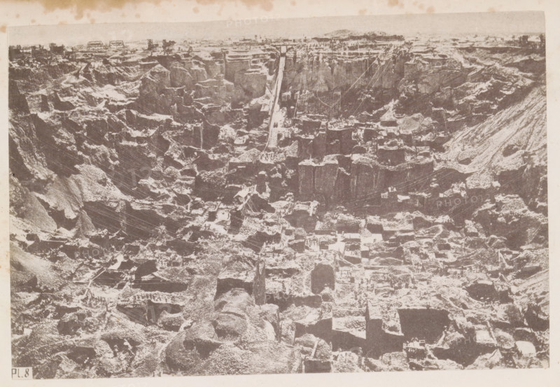 View of a diamond mine in South Africa, presumably near Kimberley, page, photomechanical print, number: 'Pl. 8', photographer: Gray Brothers (possibly), South Africa, c. 1870 - in or before 1880, paper, collotype, photogravure, height 127 mm x width 190 mm, Print in rear image section., mine, Kimberley, A high-contrast monochrome aerial view of a large excavation site showing steep terraced rock faces and irregular square and rectangular cutouts, with exposed strata creating a stepped pattern across the scene and narrow ledges and pathways between the cut blocks, scattered equipment and small structures visible on some ledges, a long straight ramp or track running from the upper edge toward the center, a few small vertical poles and thin cables spanning parts of the pit, rough rubble and loose rock piles at various levels, and a narrow strip of lighter sky and distant low land along the top edge.