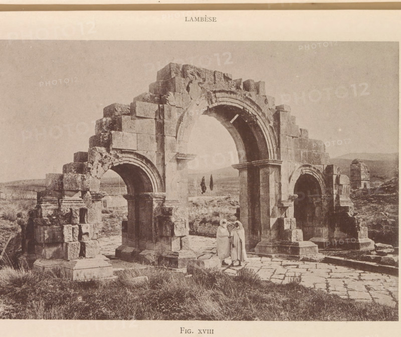 Arc de Septime Sévére, View of the arch of Septimius Severus at Lambaesis in Algeria, page, photomechanical print, number: 'Fig. XVIII', Algeria, maker: Berthaud Frères, France, c. 1884 - in or before 1894, photographer, North Africa, paper, collotype, photoprint, height 105 mm x width 151 mm, Print after page 24., triumphal arch, Lambaesis, A sepia-toned photograph showing a partially ruined stone arch structure with three large arches and weathered block masonry, standing on a paved and grassy ground, with two figures wearing long robes and head coverings standing beneath the central arch, and a distant landscape with low hills visible through and beyond the arches.