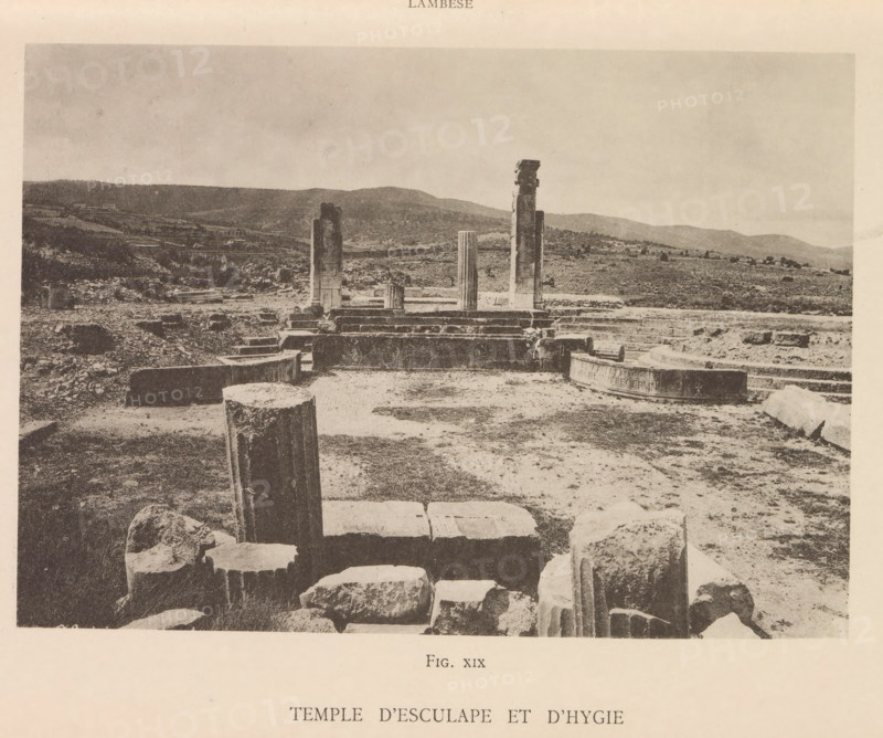 Temple d'Esculape et d'Hygie, View of the ruins of a temple of Asclepius and Hygieia at Lambaesis in Algeria, page, photomechanical print, number: 'Fig. XIX', Algeria, maker: Berthaud Frères, France, c. 1884 - in or before 1894, photographer, North Africa, paper, collotype, photoprint, height 106 mm x width 150 mm, ruin of a building, architecture, Lambaesis, A wide sepia photograph shows an archaeological site with scattered stone blocks and columns in various states of ruin across a dirt and grassy terrain, a few standing column fragments rising from a stepped platform in the midground, rounded and fluted column sections and irregular rock fragments in the foreground, low rolling hills and sparse vegetation in the background, and an expansive sky above.