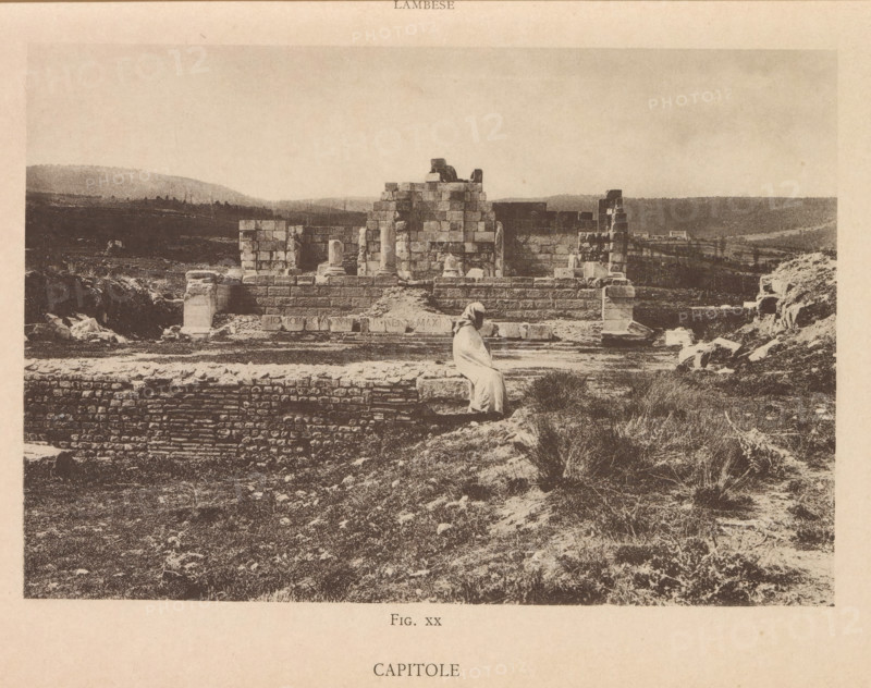Capitole, View of a Roman ruin at Lambaesis in Algeria, page, photomechanical print, number: 'Fig. XX', Algeria, maker: Berthaud Frères, France, c. 1884 - in or before 1894, photographer, North Africa, paper, collotype, photoprint, height 108 mm x width 158 mm, ruin of a building, architecture, Lambaesis, A sepia-toned photograph of an ancient stone ruin set in a grassy, uneven landscape, showing large rectangular stone blocks forming partial walls and a raised platform with a few standing columns and column fragments; a figure draped in a light-colored robe is seated on a stone ledge in the foreground facing the ruins; scattered stones and rubble lie around the structure, and the background reveals low rolling hills and sparse vegetation under a pale sky.