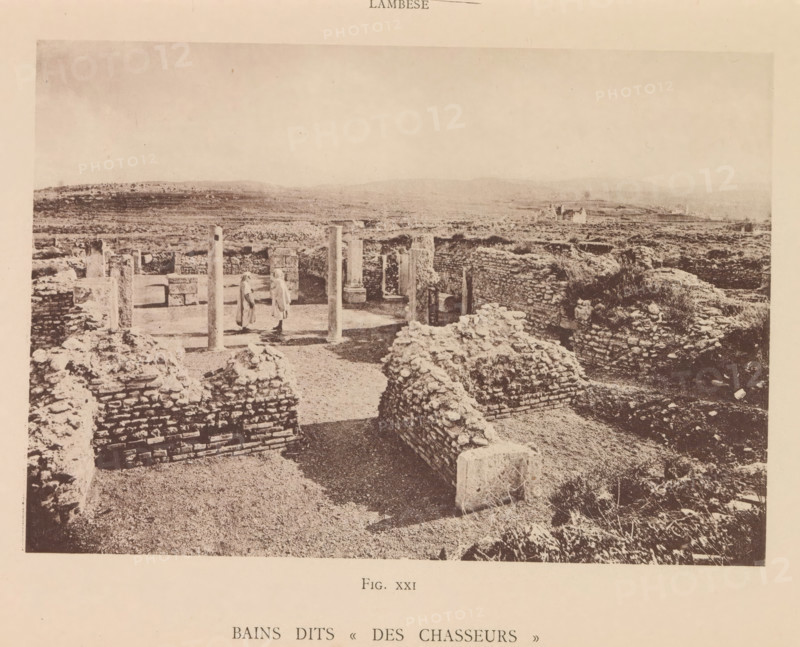 View of the ruins of a Roman bathhouse at Lambaesis in Algeria, Bains dits Des Chasseurs, page, photomechanical print, number: 'Fig. XXI', Algeria, maker: Berthaud Frères, France, c. 1884 - in or before 1894, photographer, North Africa, paper, collotype, photoprint, height 102 mm x width 147 mm, ruin of a building, architecture, public baths, Lambaesis, A wide, sepia-toned photograph shows an archaeological site with low stone walls and rubble forming rectangular enclosures across a flat, open landscape. In the foreground are rough stone mounds and short brick-like walls leading toward an inner courtyard area framed by upright columns and partial walls. Two figures wearing long garments stand near the columns within the courtyard. Beyond the ruins the land stretches to gently rolling hills under a pale sky, and scattered isolated structures appear in the distance. The image has a border of blank paper around it.