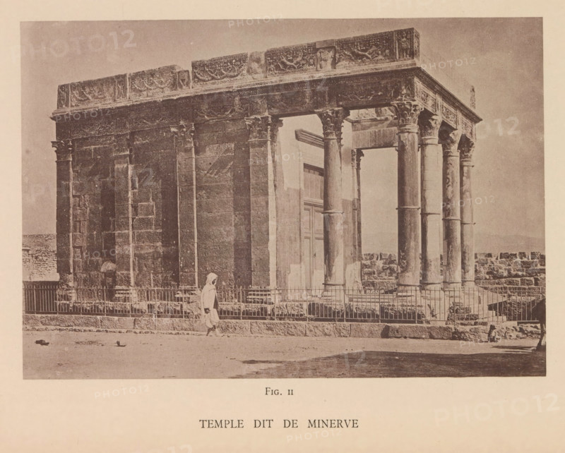 Temple dit de Minerve, View of the de Minerva temple in Tébessa, page, photomechanical print, number: 'Fig. II', Tébessa, maker: Berthaud Frères, France, c. 1884 - in or before 1894, photographer, North Africa, paper, collotype, photoprint, height 107 mm x width 157 mm, temple, shrine, Roman religion, A sepia-toned photograph of an ancient stone building with a colonnaded facade showing decorative friezes along the top and carved capitals on the columns, a gated low fence in front of the structure, an arched doorway set back between columns, a person wearing a light-colored cloak walking along the front of the fence, scattered stones and small debris on the ground in the foreground, and a low stone wall extending from the right side of the building.