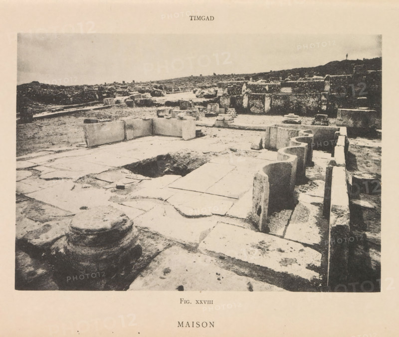 View of the Roman ruins of a house in Timgad, Maison, page, photomechanical print, number: 'Fig. XXVIII', Timgad, maker: Berthaud Frères, France, c. 1884 - in or before 1894, photographer, North Africa, paper, collotype, photoprint, height 111 mm x width 157 mm, ruin of a dwelling, house, castle, A wide, sepia-toned photograph of an archaeological ruin showing large stone slabs and blocks arranged on a flat ground, with carved stone edges and rounded stone features in the foreground. The scene includes a rectangular depression in the paved surface and several upright and fallen stone blocks scattered across the area. Along the right side there is a low stone wall with curved cutouts and a narrow trench beside it. In the background are low mounds of earth and additional ruined stone structures stretching to the horizon under a pale sky.