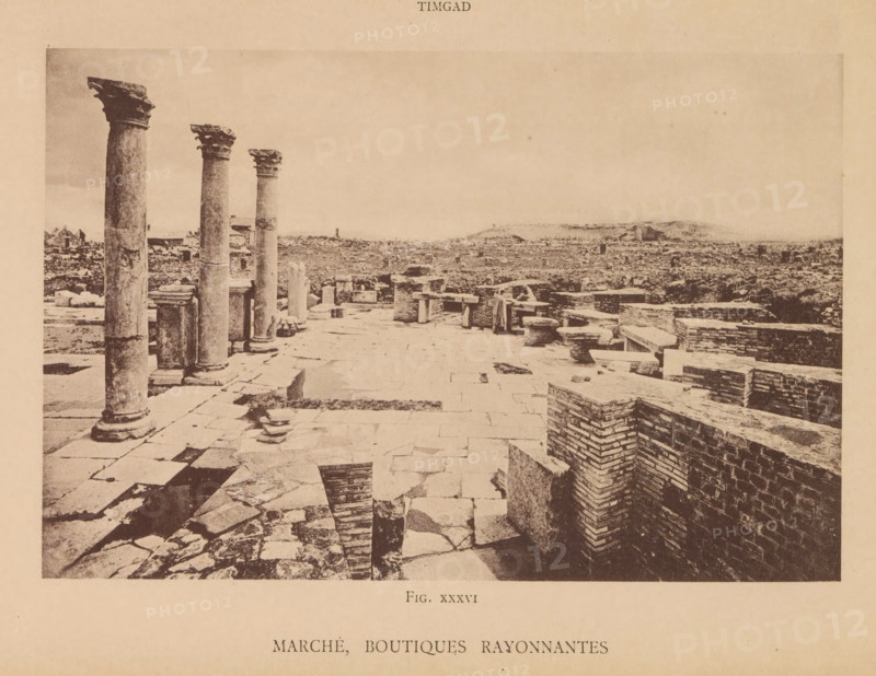 Marché, boutiques rayonnantes, View of Roman ruins of a market and shops in Timgad, page, photomechanical print, number: 'Fig. XXXVI', Timgad, maker: Berthaud Frères, France, c. 1884 - in or before 1894, photographer, North Africa, paper, collotype, photoprint, height 105 mm x width 159 mm, market-hall, landscape with ruins, A wide sepia-toned view of an ancient stone site showing a paved foreground with irregular flagstones and scattered fragments, a row of tall weathered columns with decorative capitals along the left side, low brick and stone walls forming rectangular enclosures on the right, a few broken column drums and rectangular blocks placed near the columns, and a distant flat landscape beyond the ruins under a pale sky.