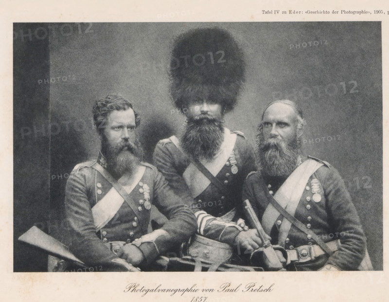 Portrait of three unknown men in uniform, page, photomechanical print, number: 'Tafel IV', photographer: Paul Pretsch, cliché maker: anonymous, c. 1900 - in or before 1905, paper, photogalvanography, height 123 mm x width 182 mm, Print after page 484., adult man, (military) uniforms, A black and white photograph of three uniformed men seated closely together, each wearing dark jackets with light diagonal sashes and rows of round buttons; the man in the center wears a very tall, furry hat and has a long full beard, the man on the left has curly hair and a thick beard and holds a long musket-like object across his lap, and the man on the right has a receding hairline with a long beard and rests one hand on a sword hilt and the other near a pouch at his waist; medals and decorative straps are visible on their chests and a drum or cylindrical object sits in front of the center figure.