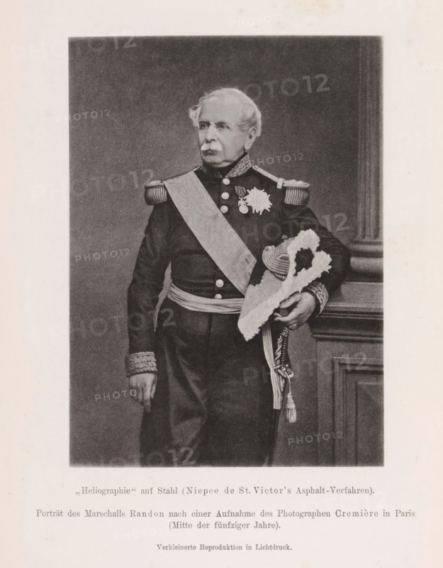 Portrait of Jacques Louis Randon, Porträt des Marschalls Randon nach einer Aufnahme des Photographen Cremière in Paris, page, photomechanical print, number: 'Tafel VI', inscription: 'Erste Proben der Generalstabskarte des kuk, 1869', photographer: Léon Cremière, Paris, cliché maker: anonymous, c. 1900 - in or before 1905, paper, collotype, photoprint, height 141 mm x width 102 mm, Print after page 484., Person, Jacques Louis Randon, commander-in-chief, general, marshal, A monochrome portrait of an older man standing in formal military dress, shown from head to mid-thigh, with light colored hair and a mustache, wearing a dark uniform with decorative shoulder epaulettes and a patterned sash across his chest, medals and a star-shaped decoration on his chest, ornate cuffs, and a sash around his waist; he holds a plumed bicorn hat with braided cords in one hand while the other hand rests at his side, and a carved wooden column or panel is visible beside him against a plain dark background, ...
