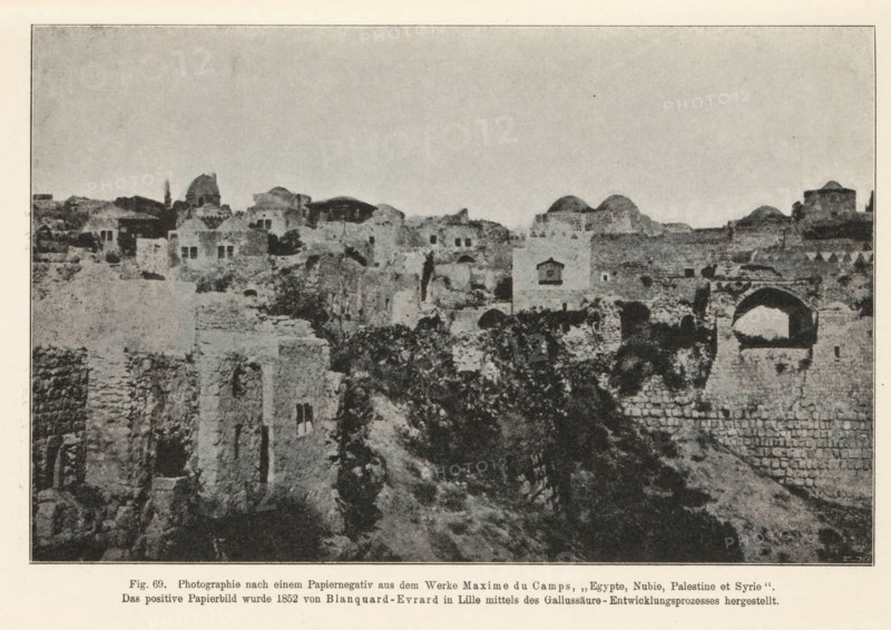 Adjutant des Fürsten Danilo von Montenegro. Nach einem Papiernegativ von P. Jovanovits, Belgrad (1850 or 1852), View of Jerusalem, page, photomechanical print, number: 'Fig. 69.', inscription: 'Photographie nach, hergestellt.', photographer: Maxime Du Camp, Jerusalem, cliché maker: anonymous, c. 1900 - in or before 1905, paper, autotypie, height 115 mm x width 118 mm, city-view in general, 'veduta', A wide monochrome photograph of an old stone settlement showing numerous ruined and intact masonry structures spread across a rocky hillside under a pale sky; visible are blocky buildings with flat and domed roofs, arched openings, small rectangular windows, and sections of crumbling walls and terraces; vegetation grows among the stones and along the slopes, and a prominent archway stands near the foreground beside stacked stone courses.