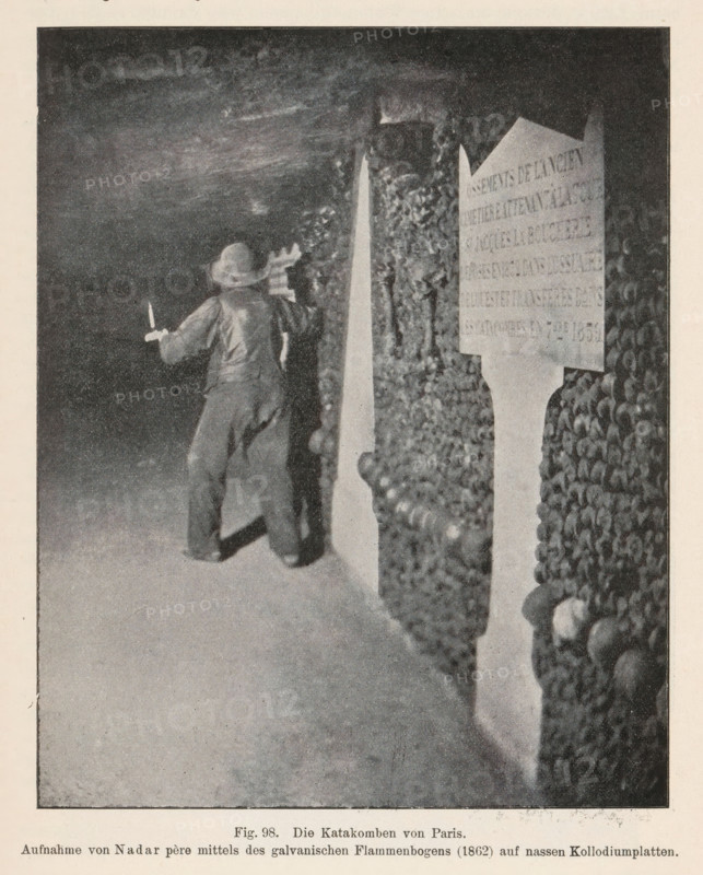 Die Catacomben von Paris, Unknown man in the Catacombs of Paris, page, photomechanical print, number: 'Fig. 98.', photographer: Félix Nadar, Catacombs of Paris, cliché maker: anonymous, c. 1900 - in or before 1905, Paris, paper, autotypie, height 134 mm x width 108 mm, adult man, catacombs, A grainy black-and-white scene showing a dim underground passage with walls lined by stacked human skulls and bones, a person wearing loose clothing and a wide-brimmed hat facing the wall and holding a candle or small torch in one hand while appearing to inspect the bones, and a tall marker or plaque positioned among the ossified walls.