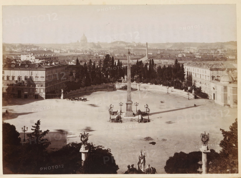View of the Piazza del Popolo with the obelisk of Ramesses II in Rome, photograph, page, Rome, c. 1850 - in or before 1860, photographer, paper, albumen print, height 97 mm x width 135 mm, square, circus, obelisk, needle, Piazza del Popolo, A wide open paved square with a tall central obelisk mounted on a stepped pedestal surrounded by decorative lamp posts and small curved benches, flanked by two symmetrical statues on pedestals at the foreground edge; the square is bordered by rows of trees and formal gardens behind which stand large multi-story buildings with rows of windows and tiled roofs, and a curved balustrade with fountains and statuary near the tree line; in the open space a few horse-drawn carriages and scattered figures are visible, and beyond the immediate buildings a distant cityscape with low buildings and a large domed structure on the horizon fills the background.