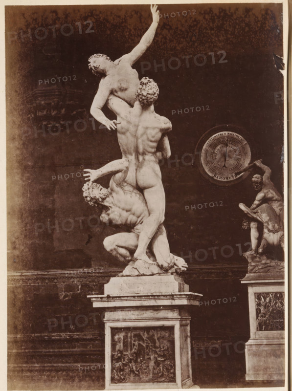 The Rape of the Sabine Virgins by Giambologna, photograph, page, Italy, c. 1850 - in or before 1860, photographer, paper, albumen print, height 132 mm x width 96 mm, piece of sculpture, reproduction of a piece of sculpture, rape of the Sabine women: they are carried off by Romulus and his men during a feast, A photograph of a large marble sculpture showing three intertwined nude figures on a rectangular pedestal, with one figure lifted high and reaching upward while another supports from below and the third crouches at the base, detailed muscular forms and curly hair visible; to the right, a smaller statue of two figures on a separate pedestal is partially visible, and a circular dial-like object is mounted on the wall behind them; the background is a dark wall with some lighter mottling and the pedestals show carved relief panels.