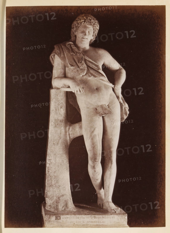 Resting Satyr by Praxiteles in the Capitoline Museums, photograph, page, Rome, c. 1850 - in or before 1860, photographer, paper, albumen print, height 134 mm x width 98 mm, piece of sculpture, reproduction of a piece of sculpture, Capitoline Museums, A photograph of a marble statue of a nude male figure leaning against a tree-like support, the figure shown from head to feet with curly hair and a relaxed facial expression, a draped cloth across one shoulder and over part of the torso, one arm resting on the support and the other hand placed near the hip, a small leaf placed over the groin, the statue standing on a rectangular base against a dark background.