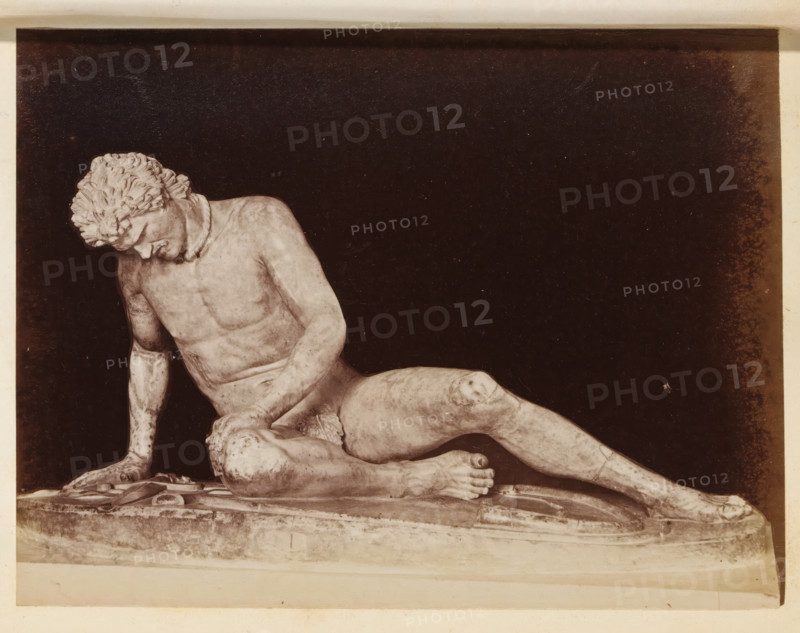 Sculpture The Dying Gaul in the Capitoline Museums, photograph, page, Rome, c. 1850 - in or before 1860, photographer, paper, albumen print, height 97 mm x width 130 mm, piece of sculpture, reproduction of a piece of sculpture, Capitoline Museums, A monochrome photograph of a reclining male marble statue set against a dark background, showing the statue's torso, head tilted downward, curly hair, one arm supporting the body with a hand on the base, and legs extended with visible muscle and anatomical detail; the statue rests on a textured rectangular plinth with visible edges and surface wear, and the image has a pale border around the dark central area.