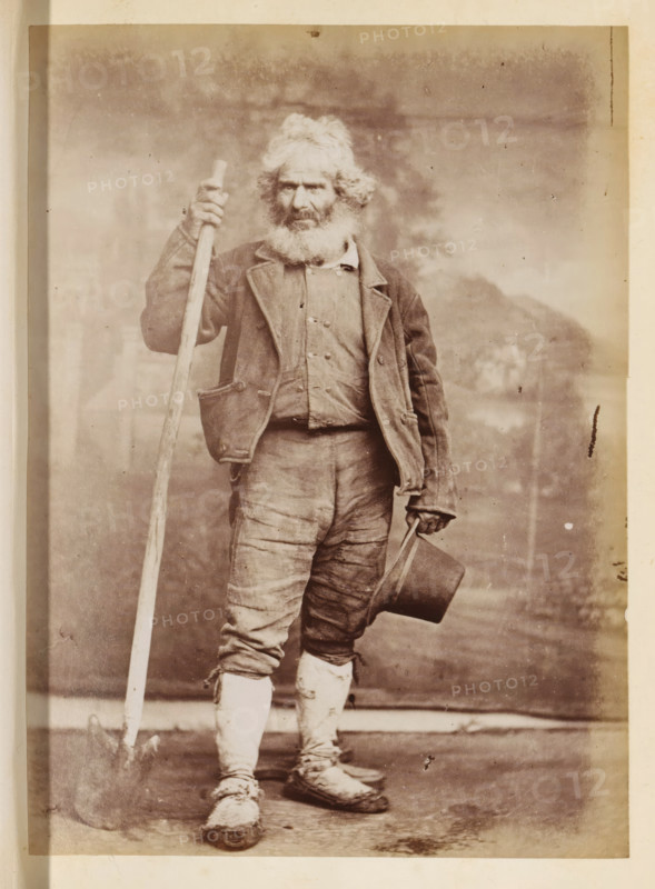 Portrait of an unknown man with a shovel and a hat in his hand, photograph, page, Rome, c. 1850 - in or before 1860, photographer, paper, albumen print, height 134 mm x width 97 mm, historical persons not known by name, adult man, A full-length portrait of an older man standing and facing forward, wearing a worn jacket over a buttoned shirt and patched trousers tucked into wrapped leggings, with a thick white beard and tousled hair; he holds a long wooden staff upright in one hand and a top hat by its brim in the other, with scuffed shoes visible at his feet against a plain backdrop.
