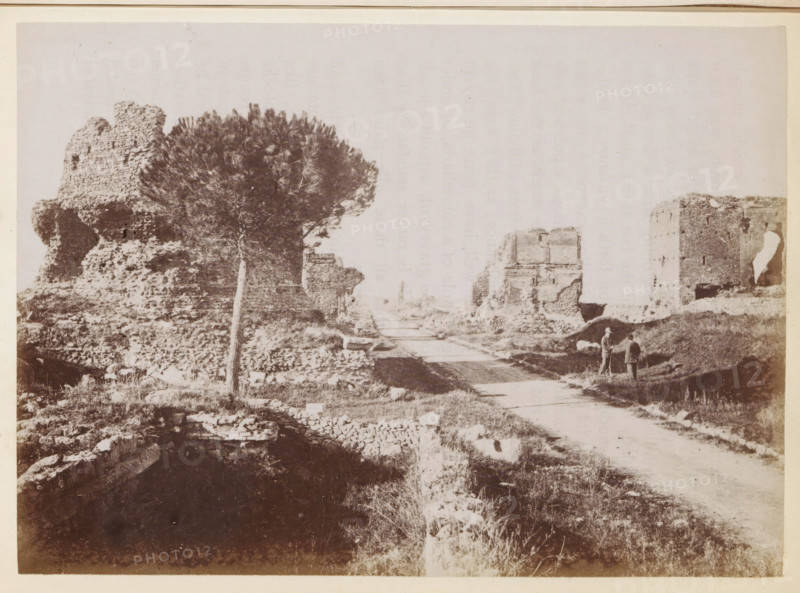 View of the Via Appia near Rome, photograph, page, Rome, c. 1850 - in or before 1860, photographer, paper, albumen print, height 97 mm x width 135 mm, street, landscape with ruins, Via Appia Antica, A wide sepia-toned scene showing rough stone ruins on both sides of a dirt road, with a large, rounded tree with dense foliage near the left ruins and scattered low vegetation across the foreground. Small sections of crumbled stone walls and masonry are visible along the roadside and in the foreground, and two small figures stand near the right edge of the road beside a mound of earth. The sky appears pale and empty above the ruins.