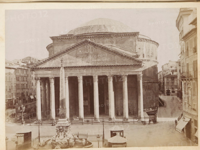 View of the Pantheon in Rome, photograph, page, Rome, c. 1850 - in or before 1860, photographer, paper, albumen print, height 99 mm x width 135 mm, church (exterior), Pantheon, A wide frontal view of a large classical building with a triangular pediment and a row of tall Corinthian columns supporting an entablature, a rounded brick drum and a large hemispherical dome rising behind it; in front of the columns stands a tall obelisk on a rectangular base set within a fenced plaza, with an ornate sculpted fountain basin visible in the foreground; flanking the plaza are multi-story buildings with shuttered windows and balconies, narrow streets, a few street lamps, a small kiosk with a canopy, and a couple of small carts and indistinct figures near the edges.