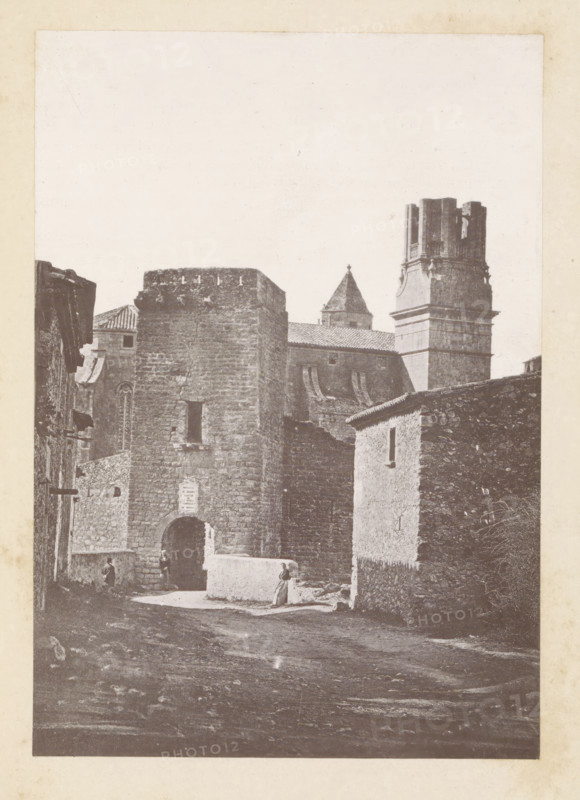 Santa Catalina gate at Torroella de Montgrí, Torroella de Montgrí, page, photomechanical print, inscription: 'Portal de Santa Catalina', c. 1873 - in or before 1883, Catalonia, Gerona (city), paper, collotype, photoprint, height 160 mm x width 111 mm, gate, entrance, A faded black-and-white photograph of a stone-built town street dominated by a tall fortified gate with an arched entrance, flanked by thick rough stone walls and adjoining buildings with small windows and tiled roofs; behind and above the gate rise a square tower and a larger ornate tower with a segmented upper section, and a pointed roof of another building is visible further back; a low whitewashed wall sits near the gate where a person stands, while another figure is near the archway; the street in the foreground is uneven and textured, and the image is bordered by a pale margin.