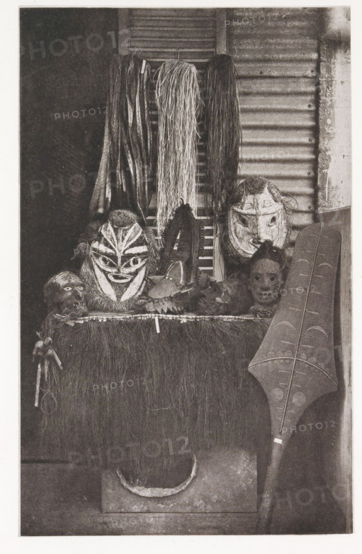 Masks and a paddle in Tahiti, Various masks, paddle, &c., page, photomechanical print, number: 'No. 23.', photographer: Henry Stuart-Wortley, Tahiti, cliché maker: Meisenbach's Autotype Company, printer (person): Meisenbach's Autotype Company, 1880, Meisenbach's Autotype Company, photographer, French Polynesia, paper, collotype, photoprint, height 168 mm x width 105 mm, mask, A vertical image showing a display of carved masks and woven materials against a textured background. At the top center several bundles of long strands hang down, varying in thickness and texture; a shutter and corrugated surface are visible behind them. Below the hanging strands are multiple carved faces and masks with distinct shapes and painted or carved facial patterns, some with hairlike material attached around them. A flat surface covered in a rough, grasslike fringe supports the masks and other small carved objects, and a carved elongated shield with patterned incisions leans against the right side. The le...
