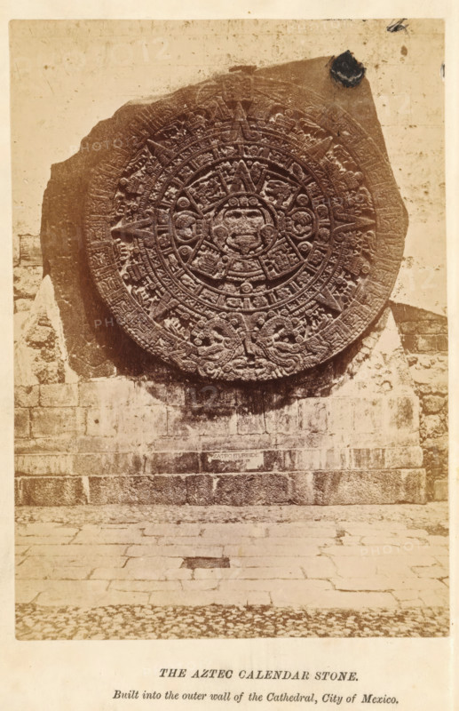 The Aztec calendar stone, photograph, page, inscription: 'Built into, City of Mexico.', Mexico City, c. 1870 - in or before 1880, photographer, North America, paper, albumen print, height 142 mm x width 100 mm, Photo on frontispiece., church (exterior), calendar, almanac, Aztecs, A large circular carved stone disk with intricate radial patterns and raised relief motifs is mounted against a rough stone wall; the disk shows concentric rings filled with geometric and figurative carvings and a central face-like figure, with serpent-like designs near the lower edge. The surrounding wall is composed of rectangular stone blocks with visible mortar lines and weathering, and a paved stone ground extends outward below the wall. The photograph has a sepia tone and shows some surface blemishes and discoloration on the wall and disk.