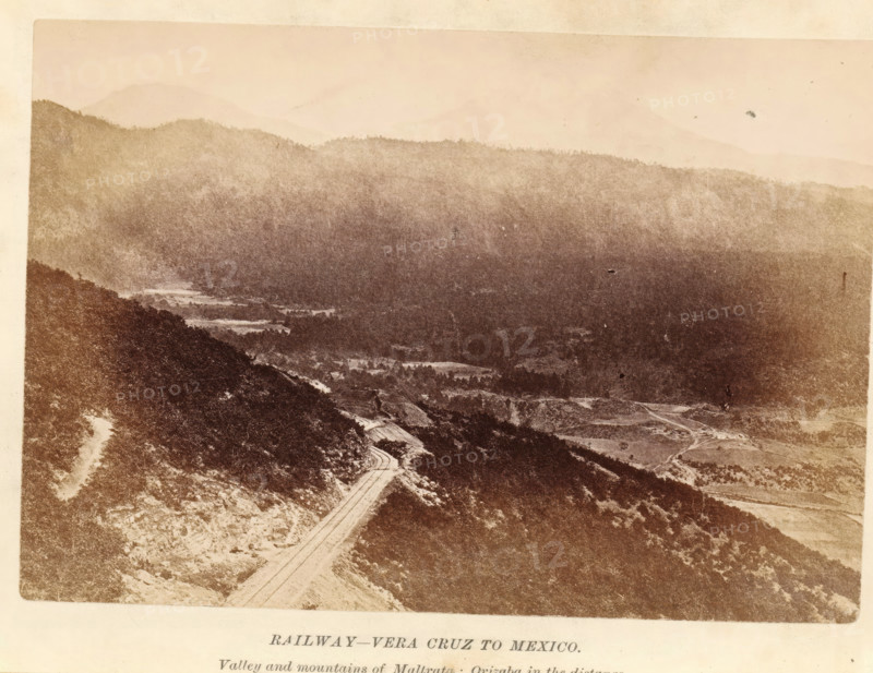 Railway-Vera Cruz to Mexico, photograph, page, inscription: 'Valley, in the distance.', Mexico, c. 1870 - in or before 1880, photographer, North America, paper, albumen print, height 97 mm x width 141 mm, railway, train, (high) hill, A sepia-toned landscape photograph showing layered hills and mountains fading into the distance under a pale sky, with darker forested ridges in the midground and a winding road cutting through a steep hillside in the foreground, alongside patches of lighter open ground and scattered trees.