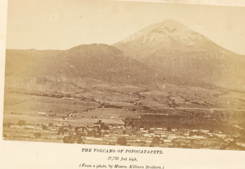 The volcano of Popocatapetl, photograph, page, inscription: '17, 726 feet high.', photographer: Kilburn, Mexico, c. 1870 - in or before 1880, North America, paper, albumen print, height 90 mm x width 154 mm, volcano, Popocatépetl, A sepia-toned photograph showing a broad landscape with a large snow-capped mountain rising behind a lower rounded hill, a patchwork of lighter and darker fields spreading across the midground, and a row of small rectangular buildings along the bottom edge, with a pale sky above.