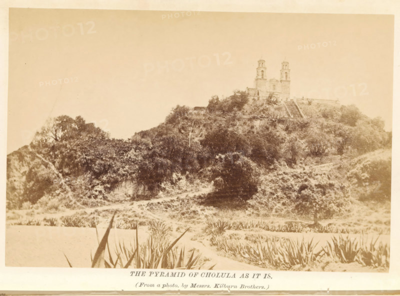 The pyramid of Cholula as it is, photograph, page, photographer: Kilburn, Mexico, c. 1870 - in or before 1880, North America, paper, albumen print, height 102 mm x width 151 mm, Photo after page 156., church (exterior), quadrangular pyramid, Great Pyramid of Cholula, Iglesia de Nuestra Señora de los Remedios, A sepia-toned photograph showing a large earthen mound covered in vegetation with a path or cleared area at its base and spiky plants in the foreground, and a stone building with twin towers situated atop the mound, stair-like terraces or steps leading up the slope, scattered trees and shrubs on the hillside, and a plain sky above.