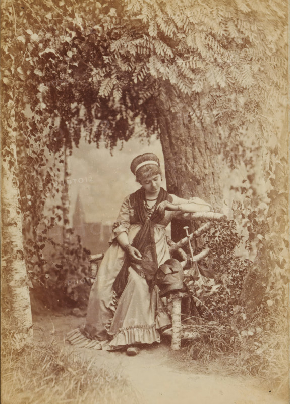 You Ring on my Finger, My gold ring, Portrait of an unknown woman sitting on a bench in front of a tree, presumably looking at a ring on her finger, Frauen-Liebe und Leben, photograph, page, number: '4.', name: 'Photog. und Verlag v. H. Hirsch, Berlin, N Christinenstr. 36 & 37', photographer: Hugo Hirsch, 1880, paper, albumen print, albumendruk, height 134 mm x width 95 mm, adult woman, bench, ring, A sepia-toned scene of a young woman leaning against a rough wooden fence beneath overhanging leafy branches, wearing a long dress with ruffled hem and layered shawls, her hair wrapped with a headband and her gaze directed downward, one arm resting on the fence and the other hand holding fabric, a hat and a walking stick hanging from the fence beside her, tree trunks and dense foliage surrounding the path behind her.