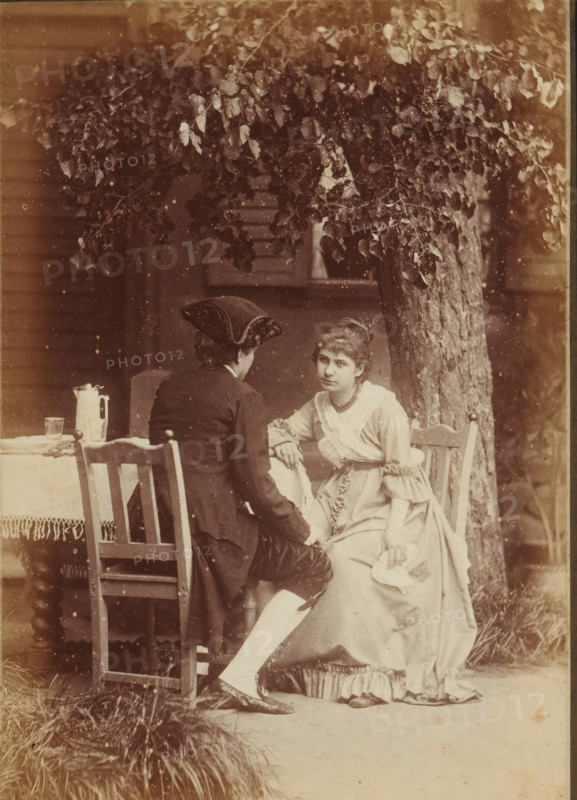 Süsser Freund, du blickest mich verwundert an, Kannst es nicht begreifen, wie ich weinen kann, Portrait of an unknown woman and man, sitting at a table in a garden, Frauen-Liebe und Leben, photograph, page, name: 'Photog. und Verlag v. H. Hirsch, Berlin, N Christinenstr. 36 & 37', number: '6.', photographer: Hugo Hirsch, 1880, paper, albumen print, albumendruk, height 134 mm x width 95 mm, garden, adult woman, adult man, historical costume, A sepia-toned scene of two people seated outdoors beneath a tree with dense leafy branches, one person facing away wearing a dark coat and a tricorne-style hat, the other facing forward wearing a light-colored dress with ruffled sleeves and a sash, the forward-facing person resting an arm on the other's knee and holding a folded cloth in the other hand, wooden chairs with simple carved backs and a small table covered with a fringed cloth visible beside them, a pitcher and a glass on the table, a tree trunk rising behind them, grass clumps at the for...