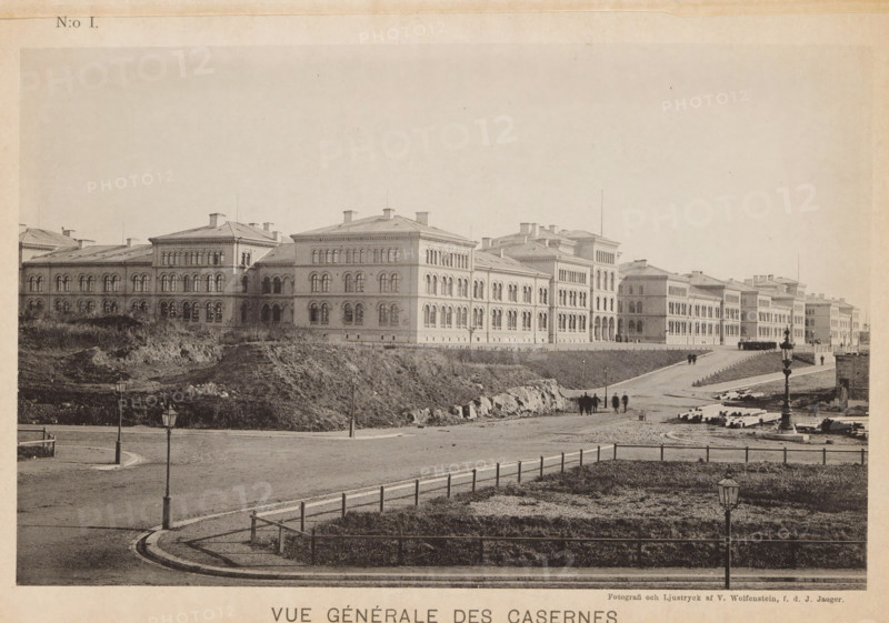 View of a barracks of the Svea livgarde at the Linnégatan in Stockholm, Vue générale des casernes, page, photomechanical print, number: 'N:o I.', maker: V. Wolfenstein, Stockholm, maker: Johannes Jaeger, c. 1888 - in or before 1891, paper, collotype, photoprint, height 120 mm x width 189 mm, barracks, A wide photographic view of a long multi-story stone building with rows of arched windows and a pitched roof stretching across the middle ground, set behind a gently sloping grassy embankment and a paved roadway; in the foreground a curved curb and fenced grassy area are visible with lamp posts lining the street, and small groups of people are walking along the road toward the buildings; scattered construction materials and rough ground appear near the embankment and the farther end of the road.