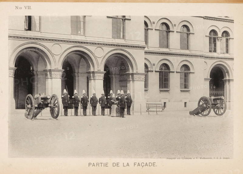 Façade of a barracks of the Svea livgarde in Stockholm with soldiers and cannons in front, Partie de la façade, page, photomechanical print, number: 'N:o VII.', maker: V. Wolfenstein, Stockholm, maker: Johannes Jaeger, c. 1888 - in or before 1891, paper, collotype, photoprint, height 117 mm x width 186 mm, the soldier, the soldier's life, firearms: cannon, façade (of house or building), A row of uniformed soldiers wearing tall helmets and dark coats stand in formation before a building facade with a series of rounded arches and ornate columns, two cannons on wooden carriages positioned on either side of the group, arched windows visible on the upper level of the building, a bench near the wall, and a paved open area in front.